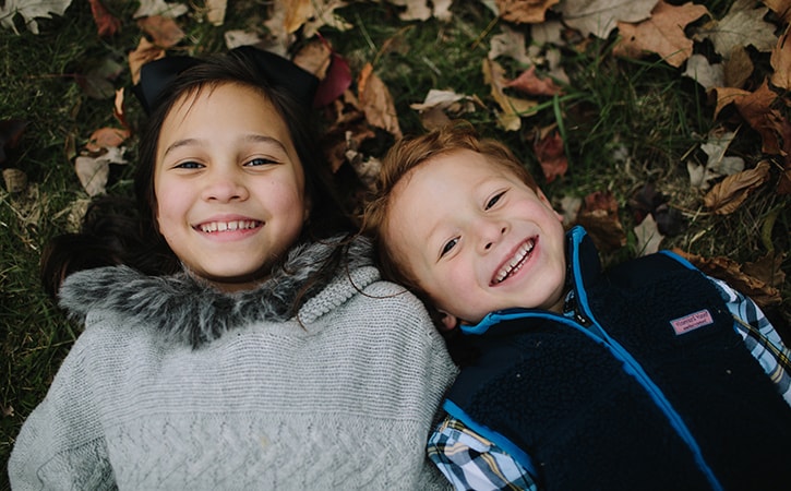 Two little boys lying on the grass and dry leaves while smiling and wearing sweaters